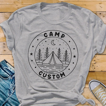 May include: A light gray short-sleeved t-shirt featuring a black circular graphic on the chest. The design includes a tent, pine trees, a crescent moon, and stars, with the text CAMP CUSTOM printed in a circular border around the illustration.