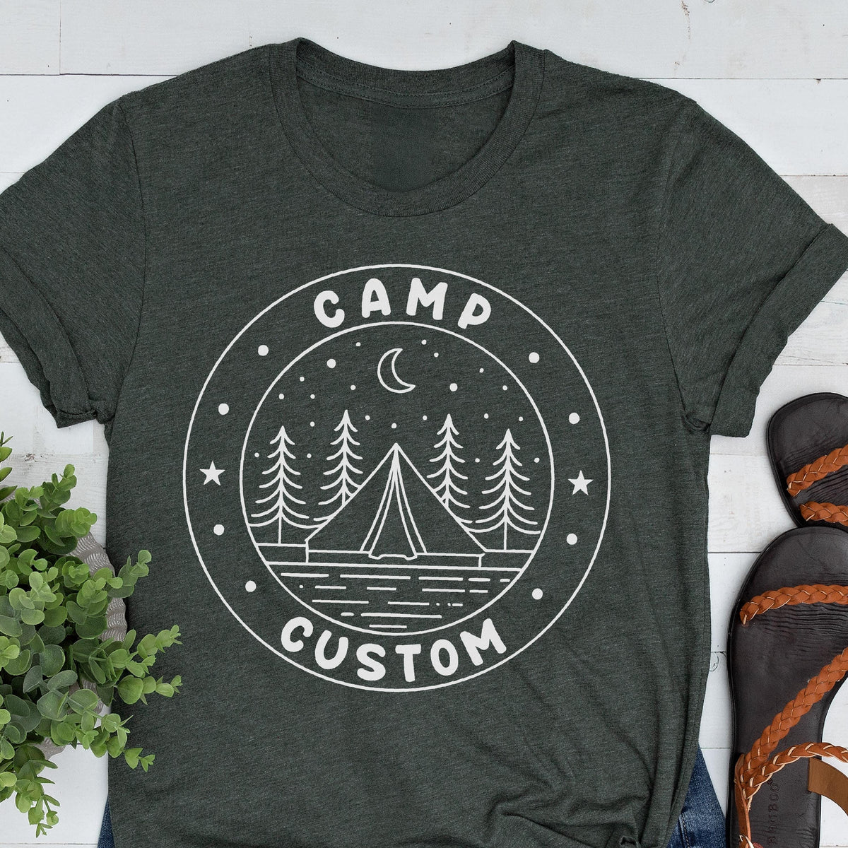 May include: A dark charcoal gray short-sleeve t-shirt featuring a white circular graphic. The design includes a tent, pine trees, a crescent moon, and stars, with the text CAMP CUSTOM printed in a bold, sans-serif font along the circular border.