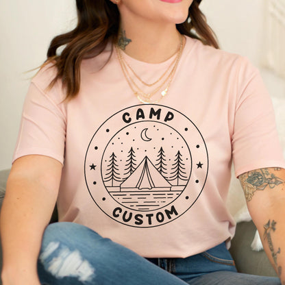 May include: A light pink short-sleeve t-shirt featuring a black circular graphic on the chest. The design includes a tent, pine trees, a crescent moon, and stars, with the text CAMP CUSTOM printed in a bold, rounded font within the circle border.
