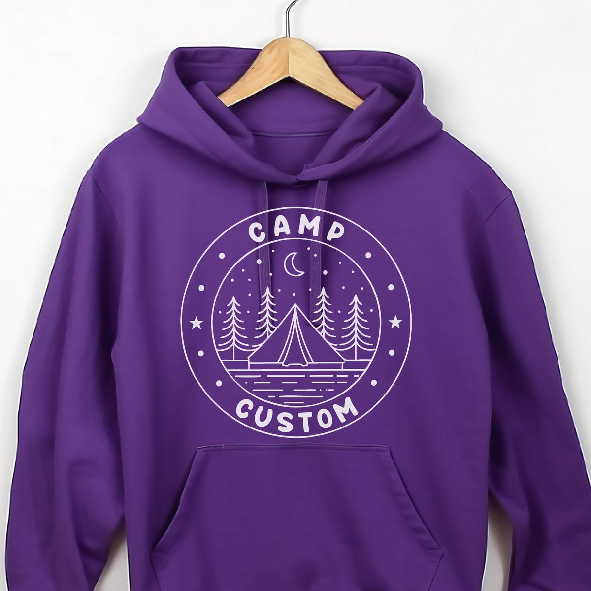 May include: A vibrant purple hooded sweatshirt featuring a white circular graphic on the chest. The design depicts a tent, pine trees, a crescent moon, and stars, with the text CAMP CUSTOM printed in a circular border around the central illustration.