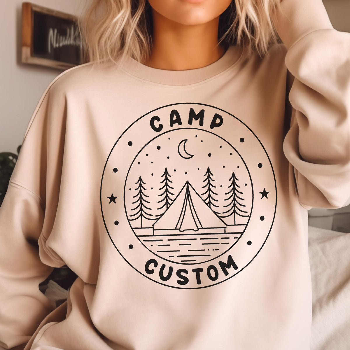 May include: A beige crewneck sweatshirt featuring a black circular graphic on the chest. The design includes a tent, pine trees, stars, and a crescent moon, with the text "CAMP" at the top and "CUSTOM" at the bottom inside the circular border.