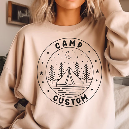 May include: A beige crewneck sweatshirt featuring a black circular graphic on the chest. The design includes a tent, pine trees, stars, and a crescent moon, with the text "CAMP" at the top and "CUSTOM" at the bottom inside the circular border.
