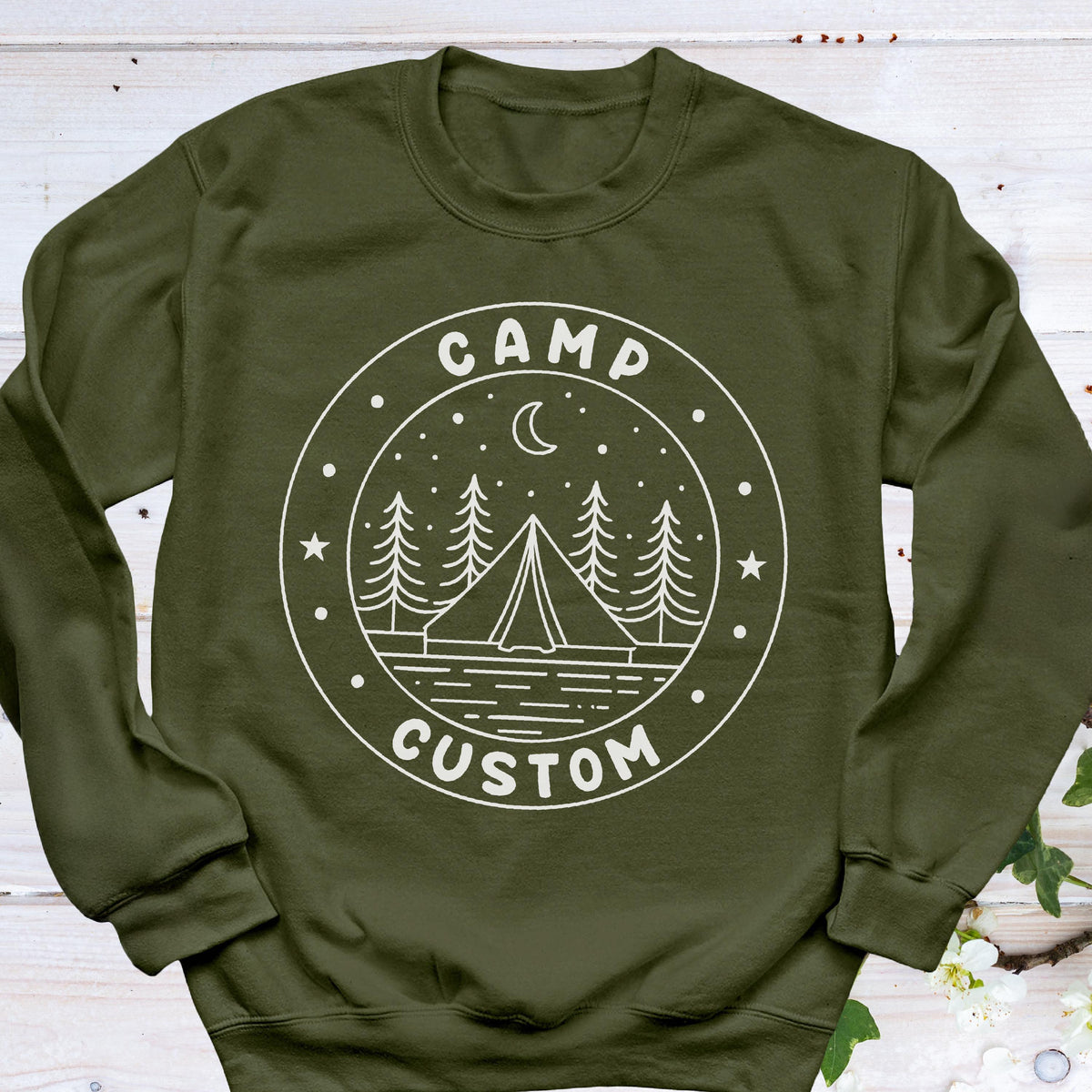 May include: An olive green crewneck sweatshirt featuring a white circular graphic on the chest. The design includes a tent, pine trees, a crescent moon, and stars, with the text "CAMP" at the top and "CUSTOM" at the bottom of the circle.