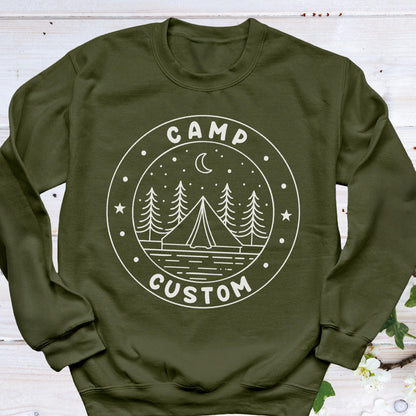 May include: An olive green crewneck sweatshirt featuring a white circular graphic on the chest. The design includes a tent, pine trees, a crescent moon, and stars, with the text "CAMP" at the top and "CUSTOM" at the bottom of the circle.