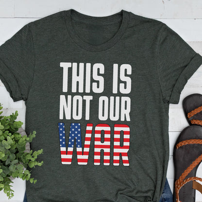 May include: Dark gray t-shirt with the text "THIS IS NOT OUR WAR" in white block letters. The word "WAR" is designed with the American flag's stars and stripes. The shirt is a casual wear item.