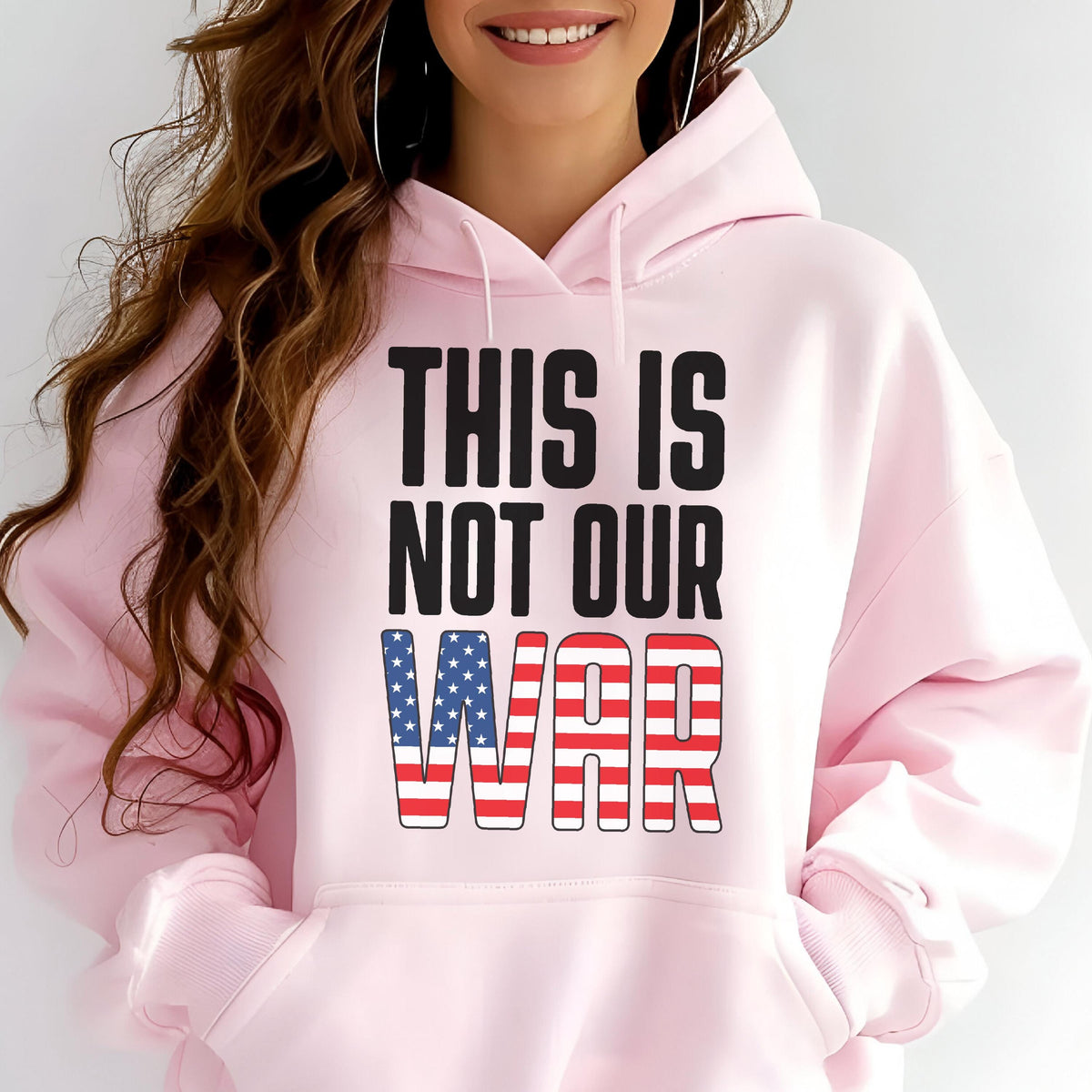 May include: A light pink hoodie with the text "THIS IS NOT OUR WAR" in black and red, with the word "WAR" designed with the American flag. The hoodie has a front pocket and a hood.