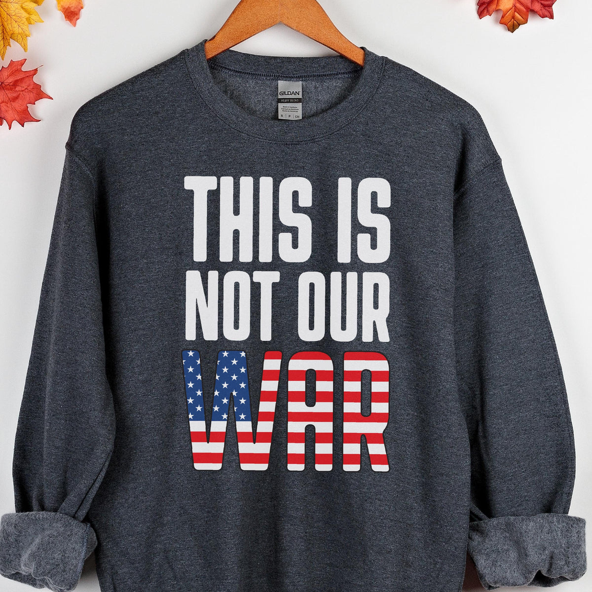 May include: A dark gray sweatshirt with the text "THIS IS NOT OUR WAR" in white block letters. The word "WAR" is designed with the American flag's stars and stripes. The sweatshirt is displayed on a wooden hanger.