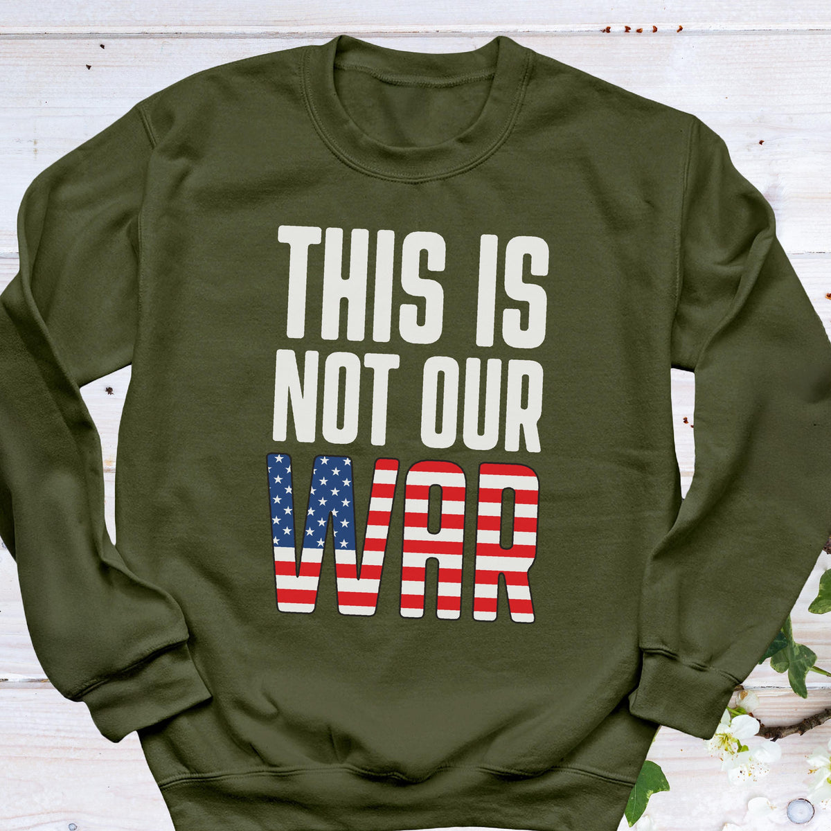 May include: An olive green sweatshirt with the text "THIS IS NOT OUR WAR" in white block letters. The word "WAR" is designed with the American flag's stars and stripes.