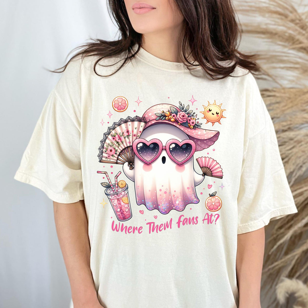 May include: Cream-colored t-shirt featuring a cartoon ghost wearing a pink hat, heart-shaped sunglasses, and holding a fan. The ghost is surrounded by illustrations of a drink, sun, and fruit. Text on the shirt reads "Where Them Fans At?"
