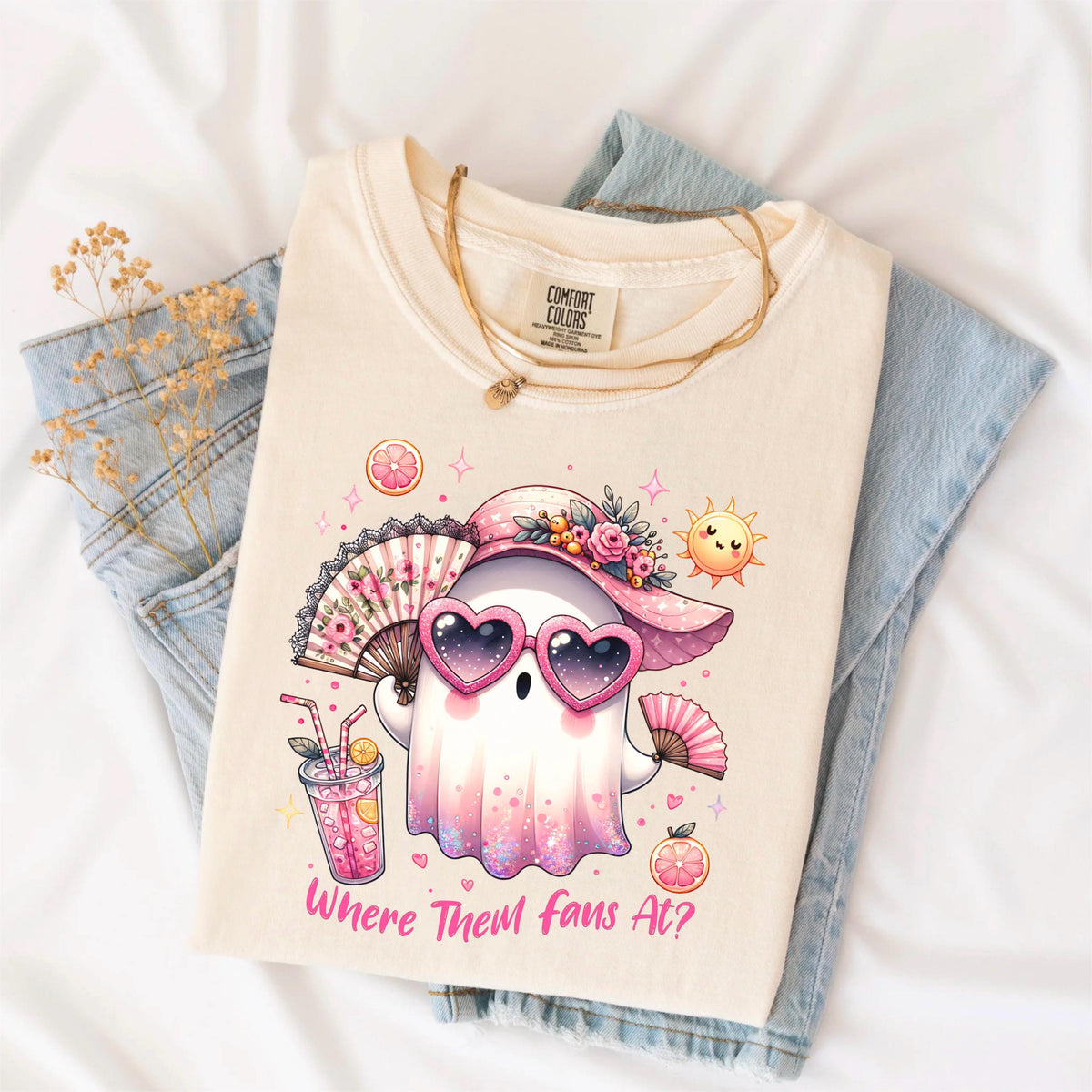 May include: Cream-colored t-shirt featuring a cartoon ghost wearing heart-shaped sunglasses and a pink hat with flowers. The ghost holds a fan and a drink, with the text "Where Them fans At?" below. The shirt is folded on top of blue jeans.