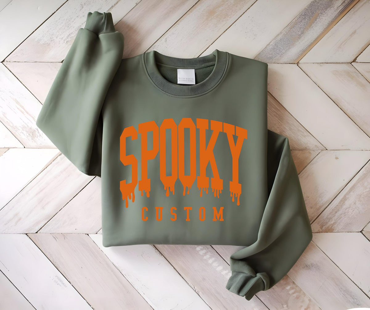 May include: An olive green crewneck sweatshirt featuring the word SPOOKY in a large, orange, dripping varsity-style font across the chest, with the word CUSTOM printed in a smaller, solid orange sans-serif font directly underneath the text.