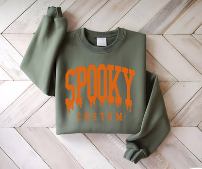 May include: An olive green crewneck sweatshirt featuring the word SPOOKY in a large, orange, dripping varsity-style font across the chest, with the word CUSTOM printed in a smaller, solid orange sans-serif font directly underneath the text.