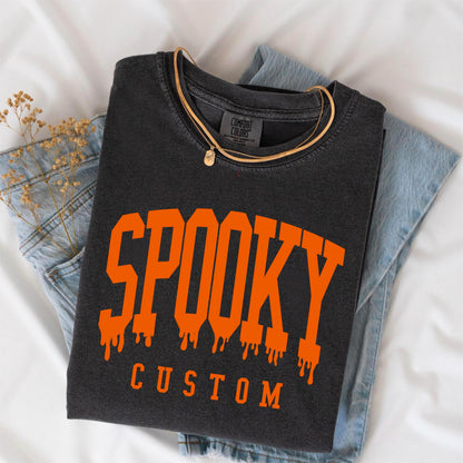 May include: A charcoal gray crewneck sweatshirt featuring the word SPOOKY in large, orange, dripping-style varsity lettering, with the word CUSTOM printed in smaller orange block letters underneath. The garment has a Comfort Colors brand label.