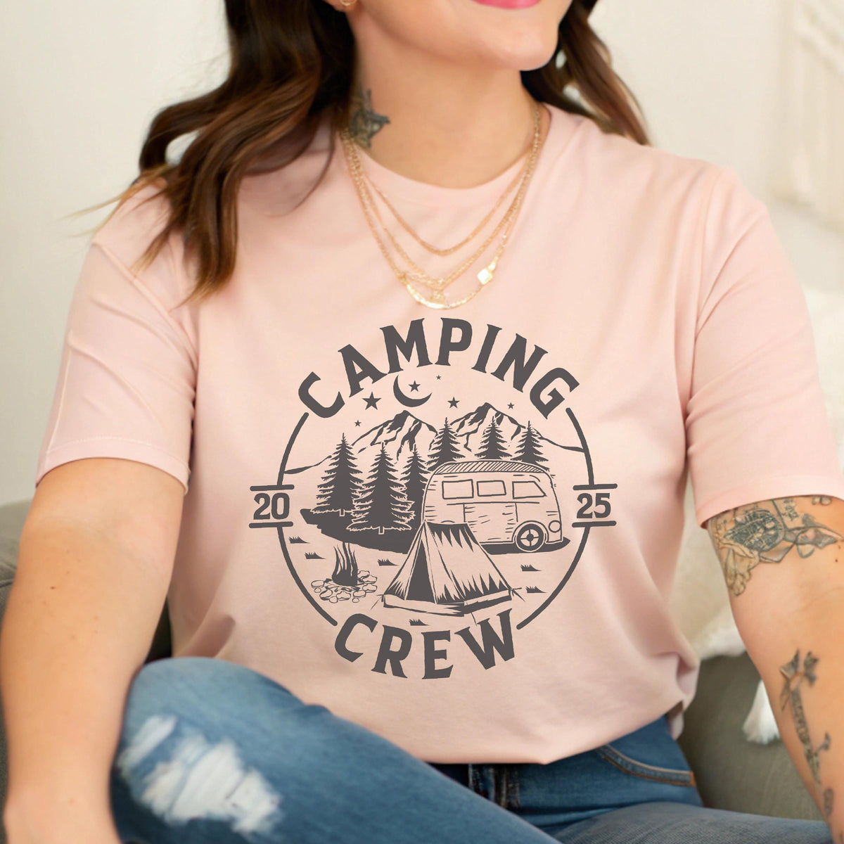May include: A light pink short-sleeve t-shirt featuring a dark gray circular graphic with the text CAMPING CREW, the years 20 and 25, a mountain range, pine trees, a crescent moon, stars, a camper van, and a tent with a small campfire.