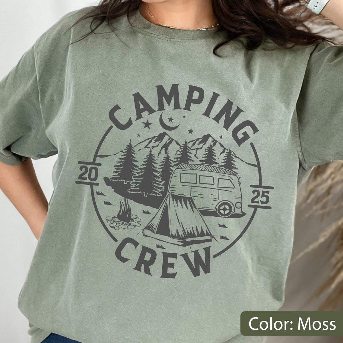 May include: A moss green short-sleeve t-shirt featuring a dark gray circular graphic. The design includes a camper van, a tent, pine trees, stars, a crescent moon, and the text "CAMPING CREW" with the years "20" and "25" on the sides.