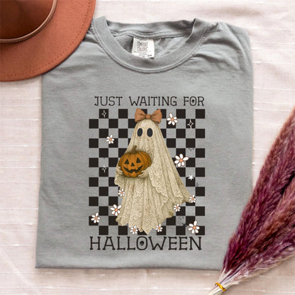 May include: A gray Comfort Colors t-shirt featuring a Halloween-themed graphic. The graphic includes a ghost holding a jack-o'-lantern, a checkered background, and the text "JUST WAITING FOR HALLOWEEN".