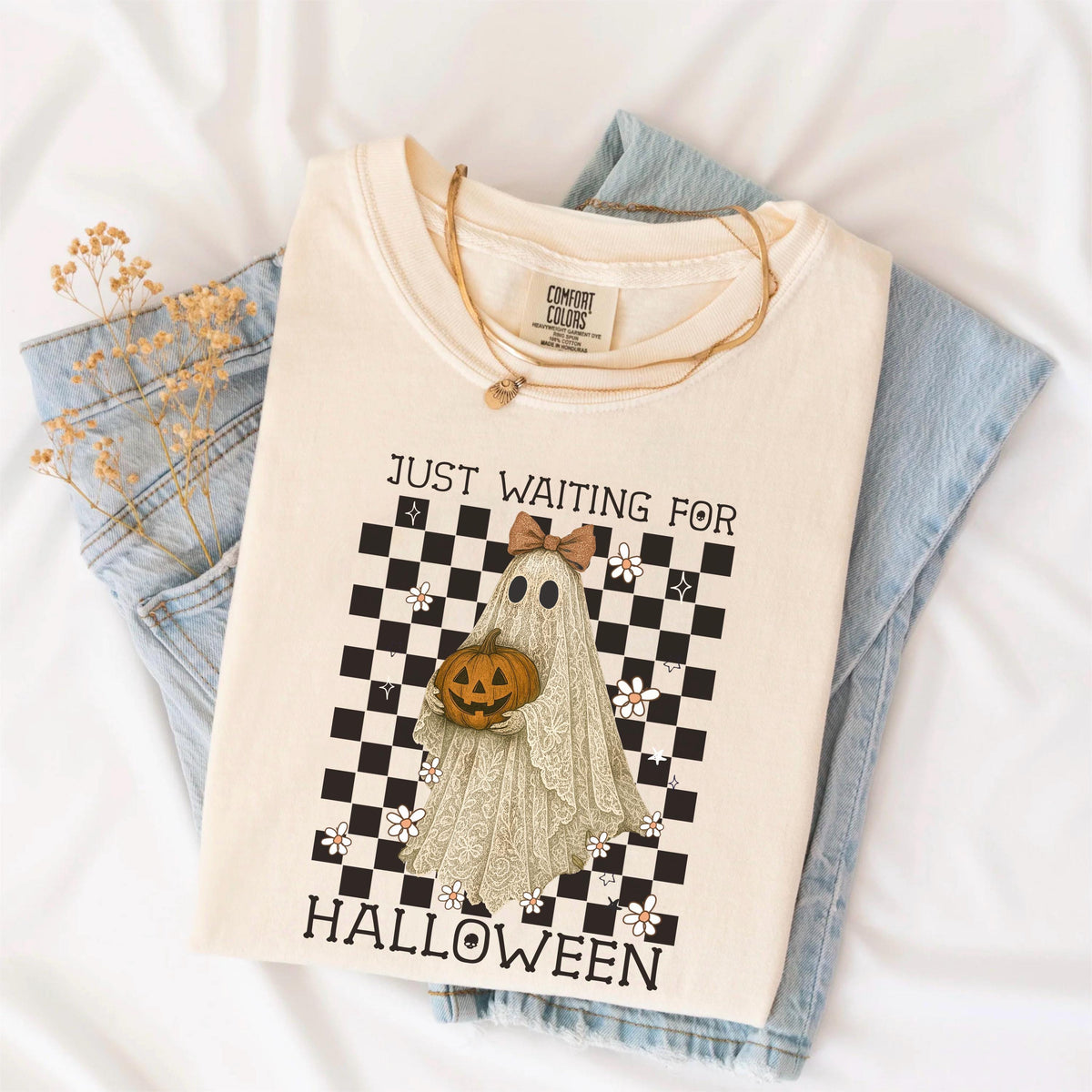 May include: Cream-colored t-shirt with the text "JUST WAITING FOR HALLOWEEN". The shirt features a ghost holding a pumpkin, set against a black and white checkered background with floral accents. A gold necklace rests on the shirt.