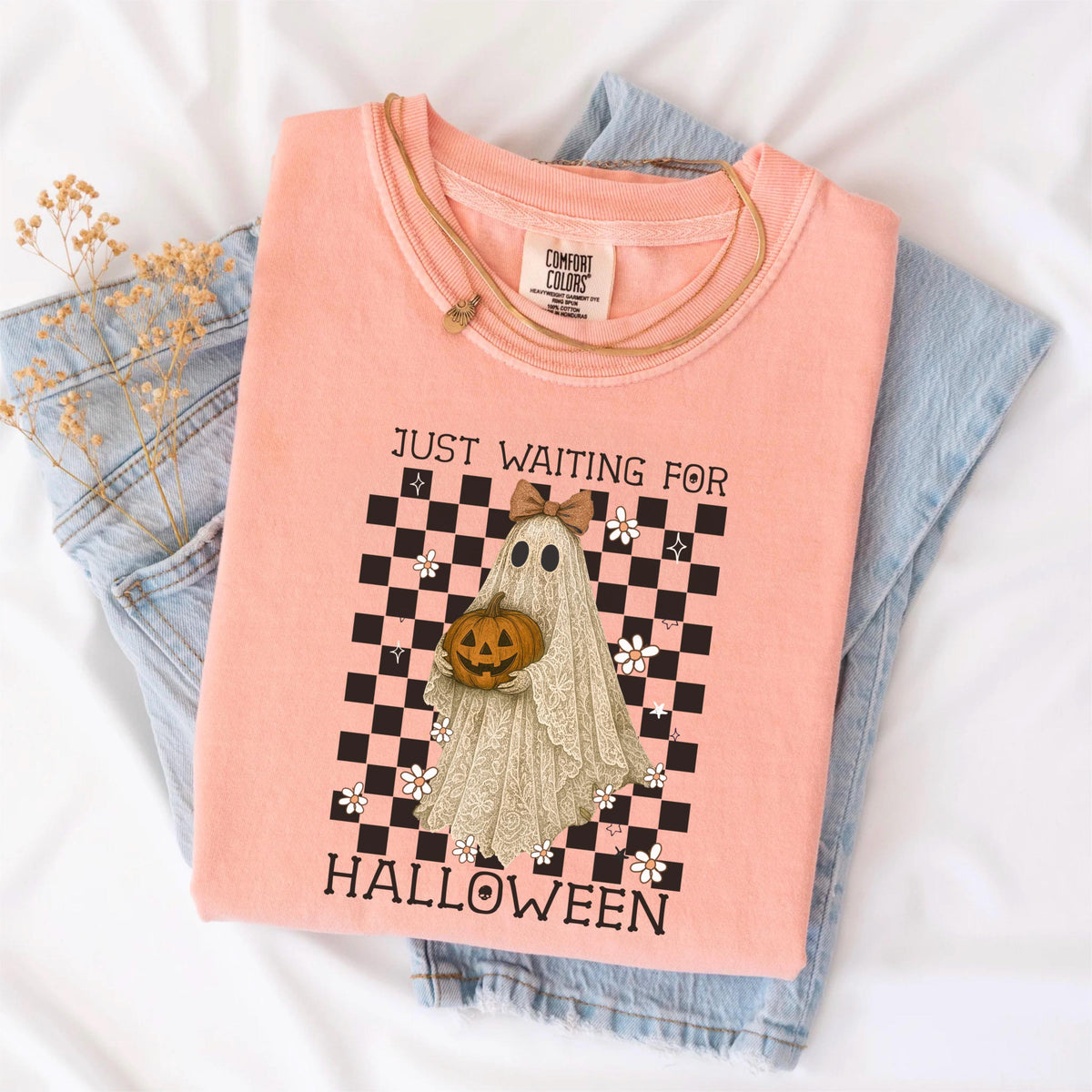May include: A coral-colored t-shirt with the text "JUST WAITING FOR HALLOWEEN" and a graphic of a ghost holding a pumpkin. The ghost is in front of a black and white checkered background. A gold necklace and blue jeans are also in the image.