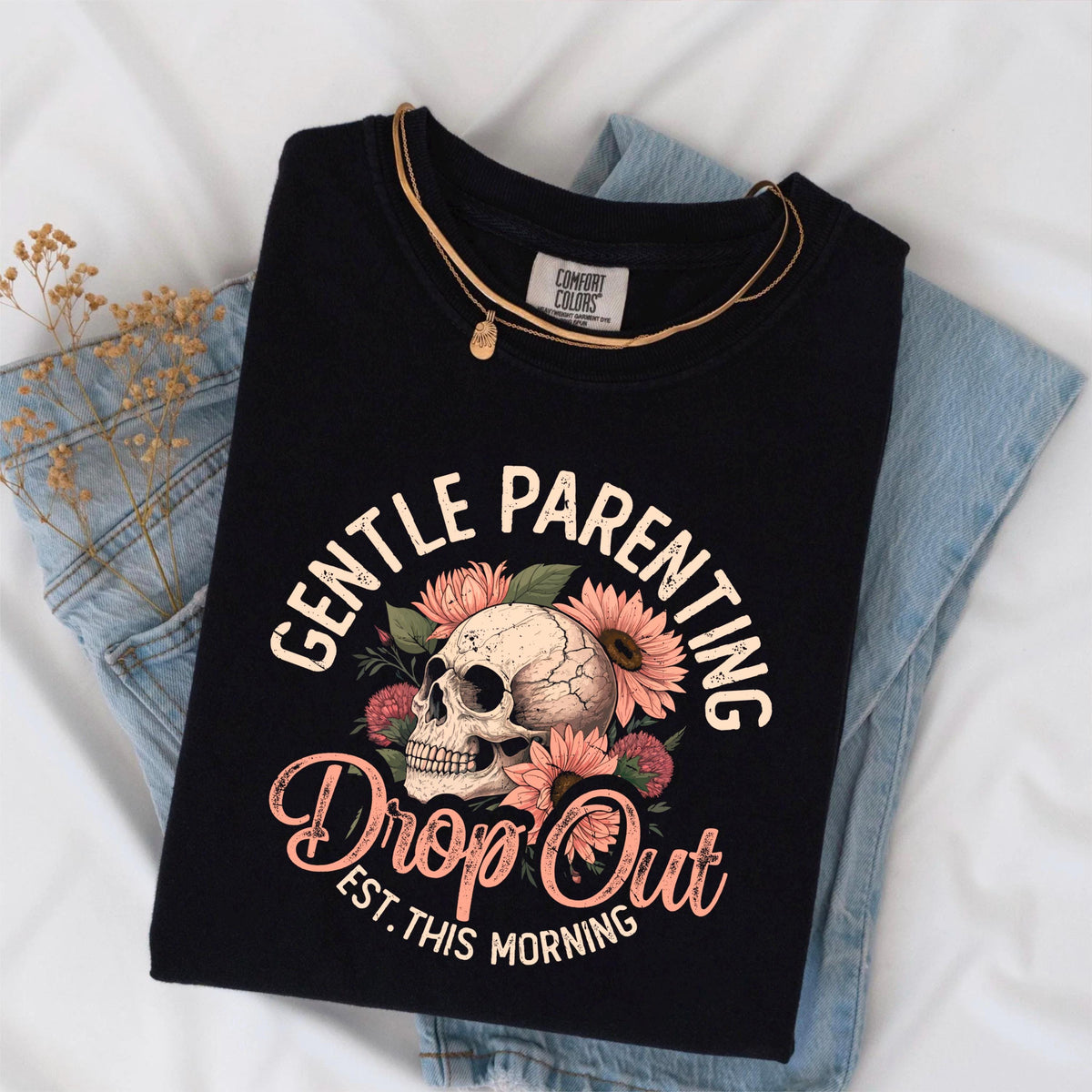 May include: Black t-shirt with the words "GENTLE PARENTING" and "Drop Out Est. This Morning" in a floral design with a skull. The shirt is folded on top of blue jeans and a gold necklace.