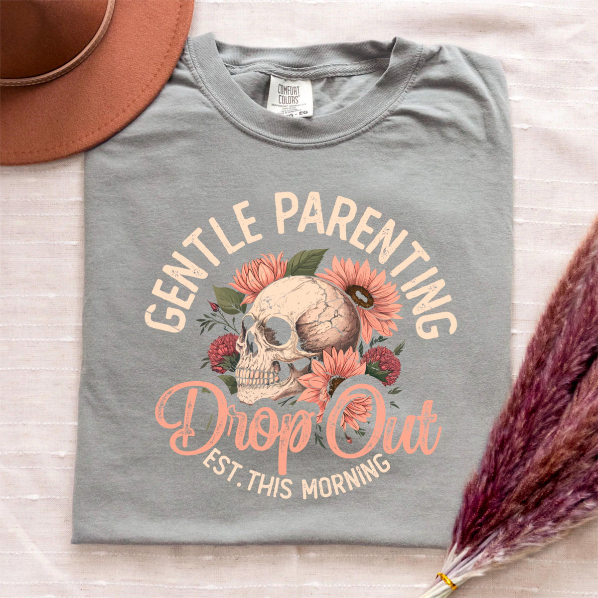 May include: A gray Comfort Colors t-shirt with the words "GENTLE PARENTING" arched above a skull surrounded by flowers. Below the skull, the words "Drop Out" and "Est. This Morning" are printed in a cursive font. A brown hat is in the upper left corner.
