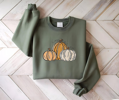 May include: Olive green sweatshirt featuring a pumpkin graphic. The graphic includes three pumpkins in shades of orange and white with black line art details. The sweatshirt has a crew neck and long sleeves.