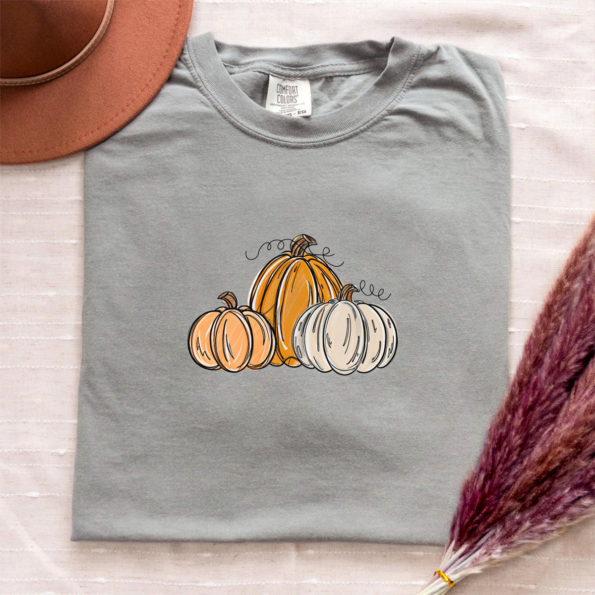 May include: A gray Comfort Colors t-shirt featuring a fall-themed design. The design includes three pumpkins in shades of orange and white, with black line art details. A brown hat is visible in the upper left corner.