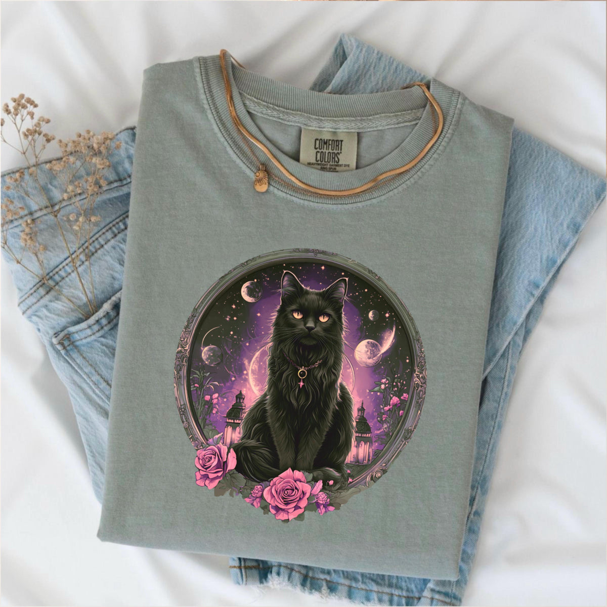 May include: A sage green Comfort Colors t-shirt featuring a detailed graphic of a black cat with glowing orange eyes, set within a circular frame with a celestial background. The cat is adorned with a necklace and surrounded by pink roses.