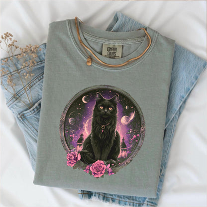 May include: A sage green Comfort Colors t-shirt featuring a detailed graphic of a black cat with glowing orange eyes, set within a circular frame with a celestial background. The cat is adorned with a necklace and surrounded by pink roses.