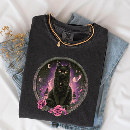 May include: Dark gray Comfort Colors t-shirt featuring a black cat illustration within a circular frame. The cat has orange eyes and is surrounded by pink roses and a celestial background. A gold necklace rests on the shirt.