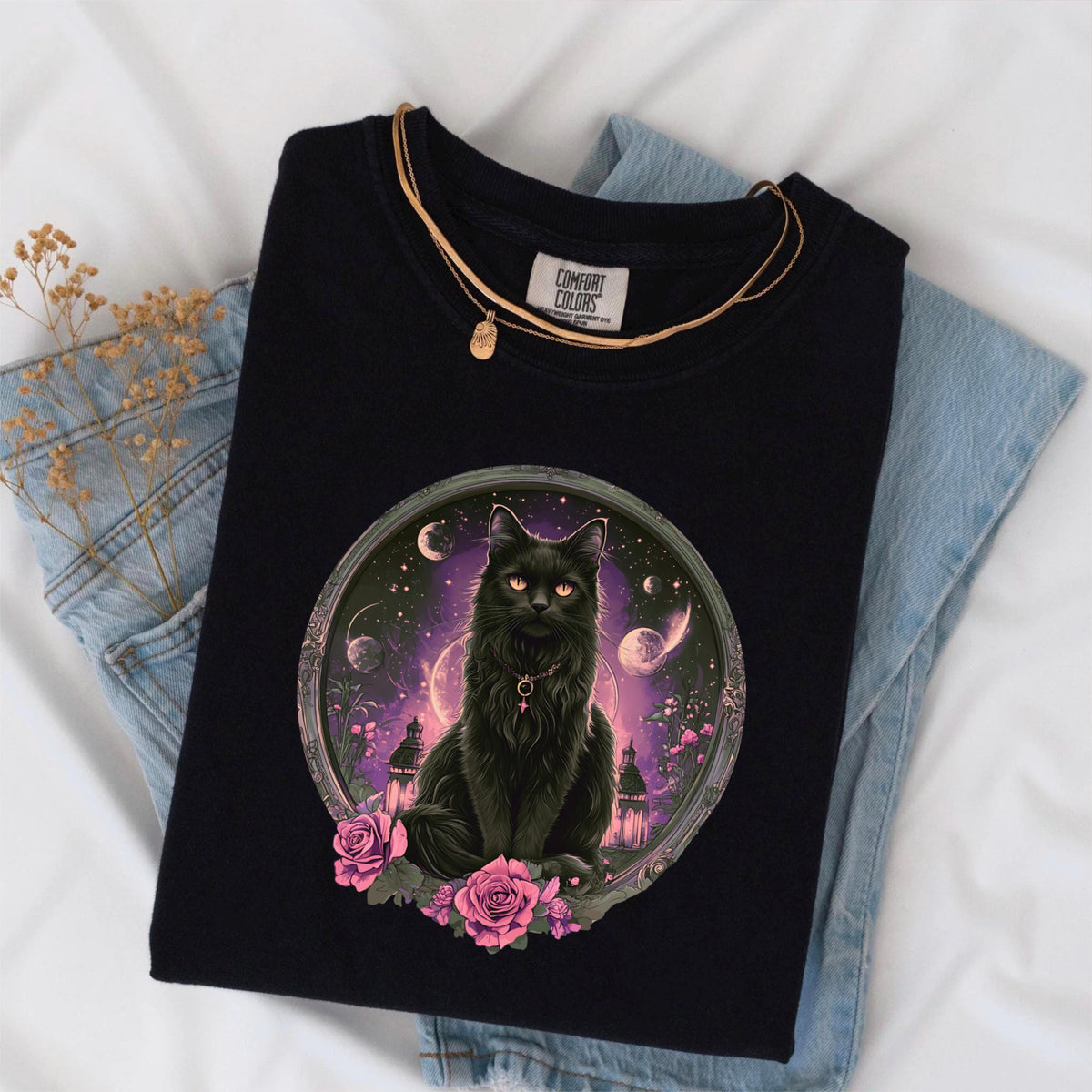 May include: Black Comfort Colors t-shirt featuring a detailed illustration of a black cat with orange eyes, set within a circular frame with a celestial background. Pink roses adorn the bottom. A gold necklace and the Comfort Colors tag are visible.