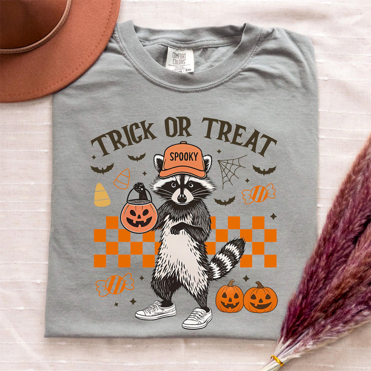 May include: A gray t-shirt with a Halloween-themed graphic. The design features a raccoon wearing an orange hat that says "Spooky" and holding a jack-o'-lantern. The text "Trick or Treat" is above the raccoon, with bats, candy corn, and pumpkins.