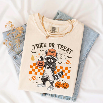 May include: Cream-colored t-shirt with a Halloween-themed graphic. The design features a raccoon wearing an orange cap and holding a jack-o'-lantern. The text "Trick or Treat" is above the raccoon, with other Halloween elements.
