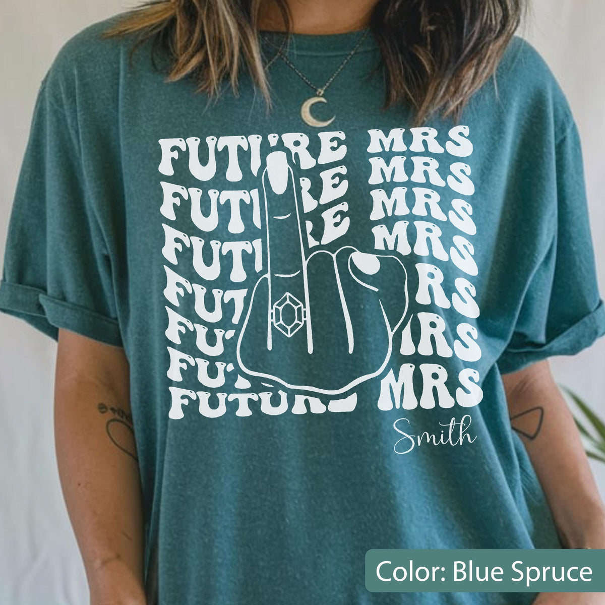May include: A blue spruce t-shirt featuring a white graphic of a hand with a ring on the middle finger. The text "FUTURE MRS" is repeated in a wavy pattern behind the hand, and the word "Smith" is printed in a script font at the bottom.