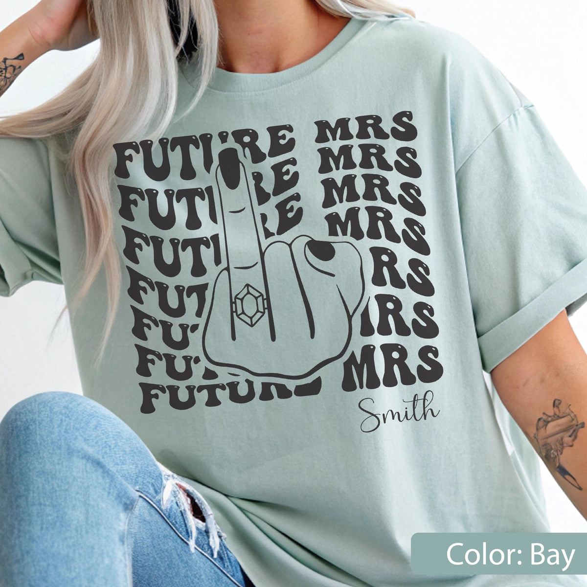 May include: A light sage green short-sleeve t-shirt featuring a black graphic of a hand making a middle finger gesture with a diamond ring on the finger. The text "FUTURE" and "MRS" is repeated in a wavy pattern, with "Smith" written below.