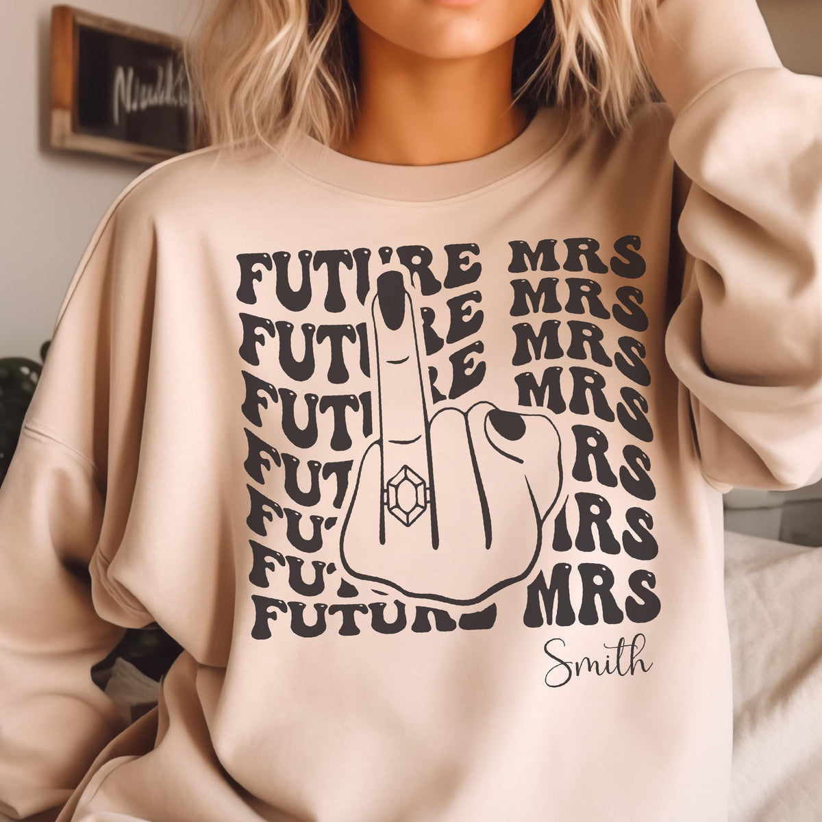 May include: Beige sweatshirt with a black graphic. The graphic features a hand with a ring on the finger, the words "FUTURE MRS" repeated, and the last name "Smith". The sweatshirt is a casual style.