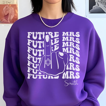 May include: A purple sweatshirt with the text "FUTURE MRS" repeated in a stacked pattern, with a graphic of a hand giving the middle finger, wearing a ring. The name "Smith" is written below the graphic.