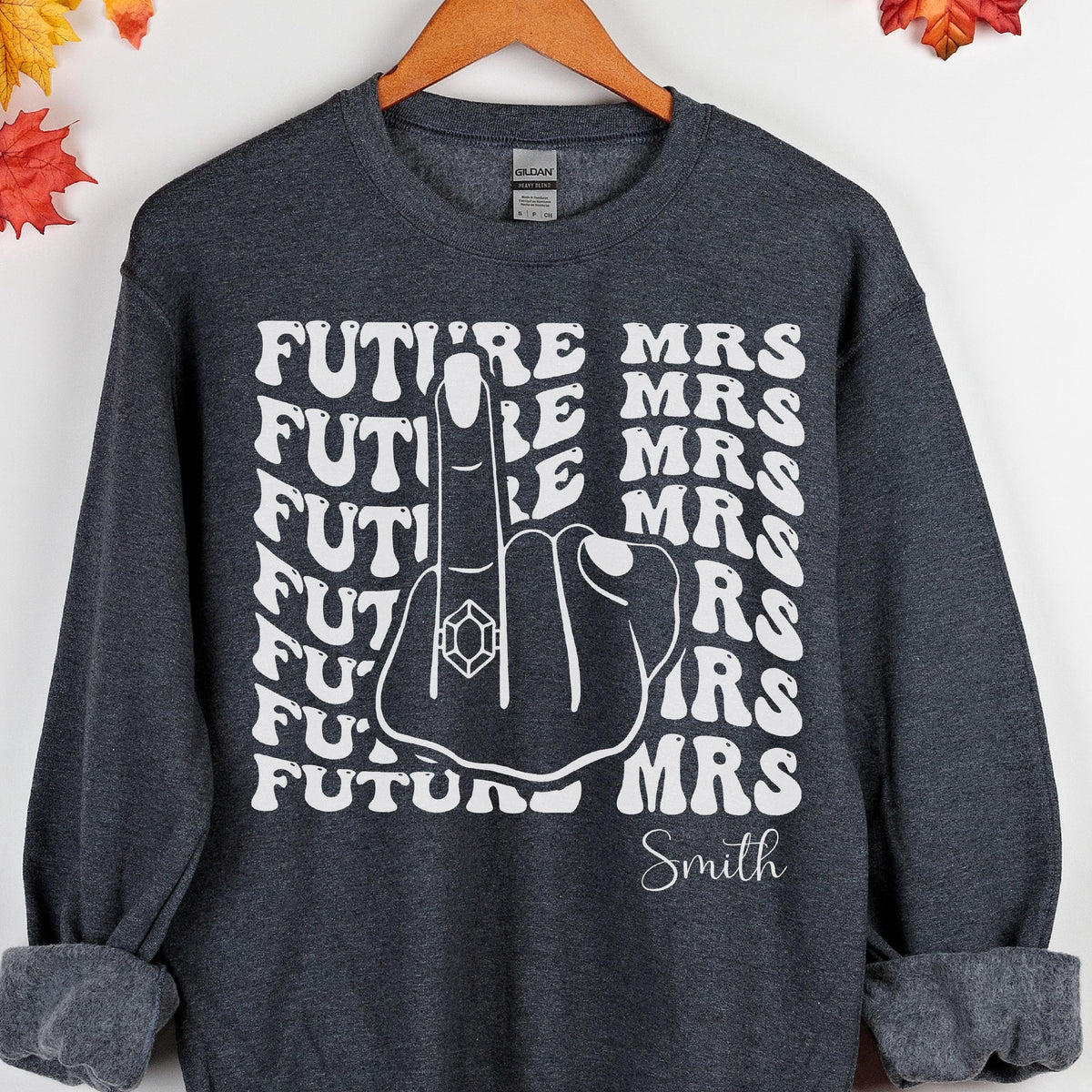 May include: Dark gray sweatshirt with white text that reads "FUTURE MRS" and "Smith". The design features a hand with a ring on the middle finger. A fun and casual bridal shower or engagement gift.