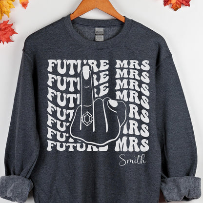 May include: Dark gray sweatshirt with white text that reads "FUTURE MRS" and "Smith". The design features a hand with a ring on the middle finger. A fun and casual bridal shower or engagement gift.