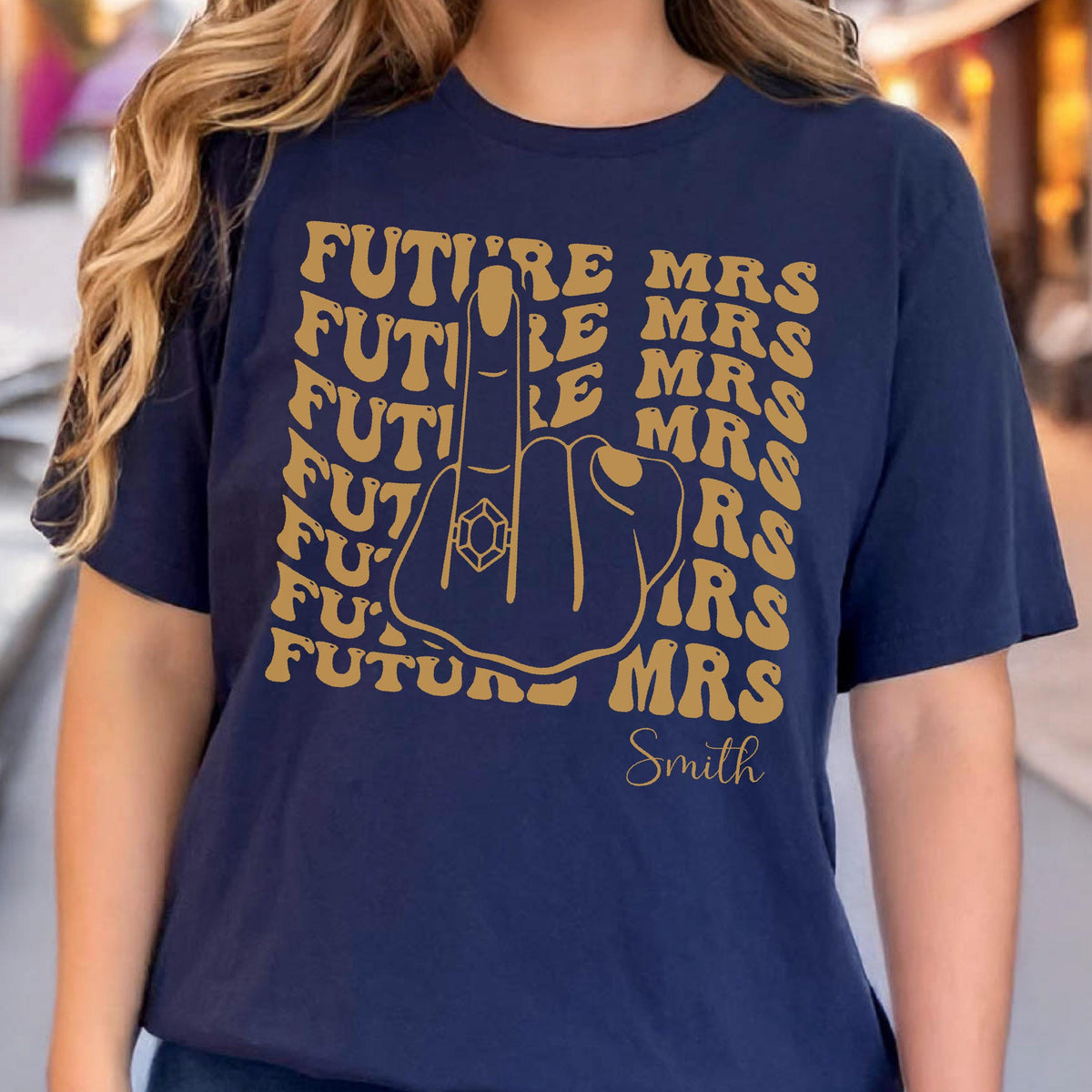 May include: A navy blue t-shirt featuring a gold graphic of a hand with a ring on the middle finger. The text "FUTURE MRS" is repeated in a wavy pattern around the hand, and the word "Smith" is printed in a script font at the bottom right.