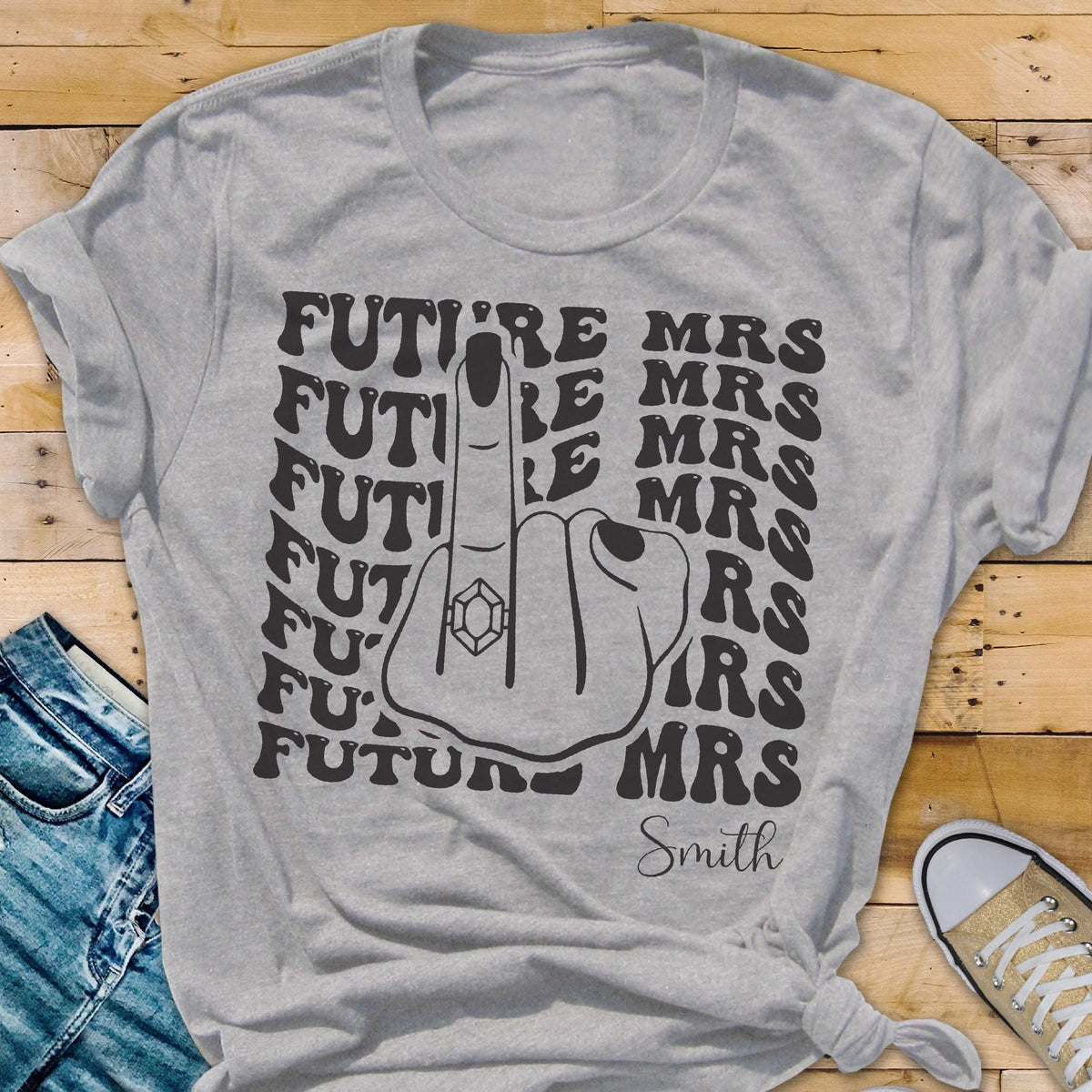 May include: A light gray t-shirt featuring a black graphic of a hand making a middle finger gesture with a diamond ring on the finger. The text "FUTURE MRS" is repeated in a wavy pattern behind the hand, and the word "Smith" is printed below.