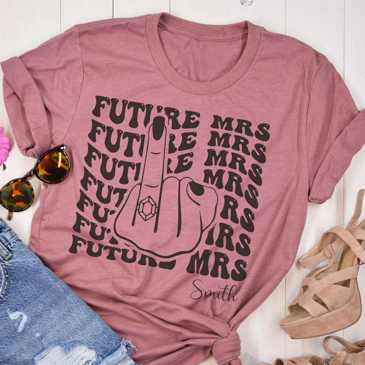 May include: A dusty rose short-sleeved t-shirt featuring a black graphic of a hand with a ring on the middle finger. The text "FUTURE MRS" is repeated in a wavy pattern, and the name "Smith" is printed in a cursive script at the bottom.