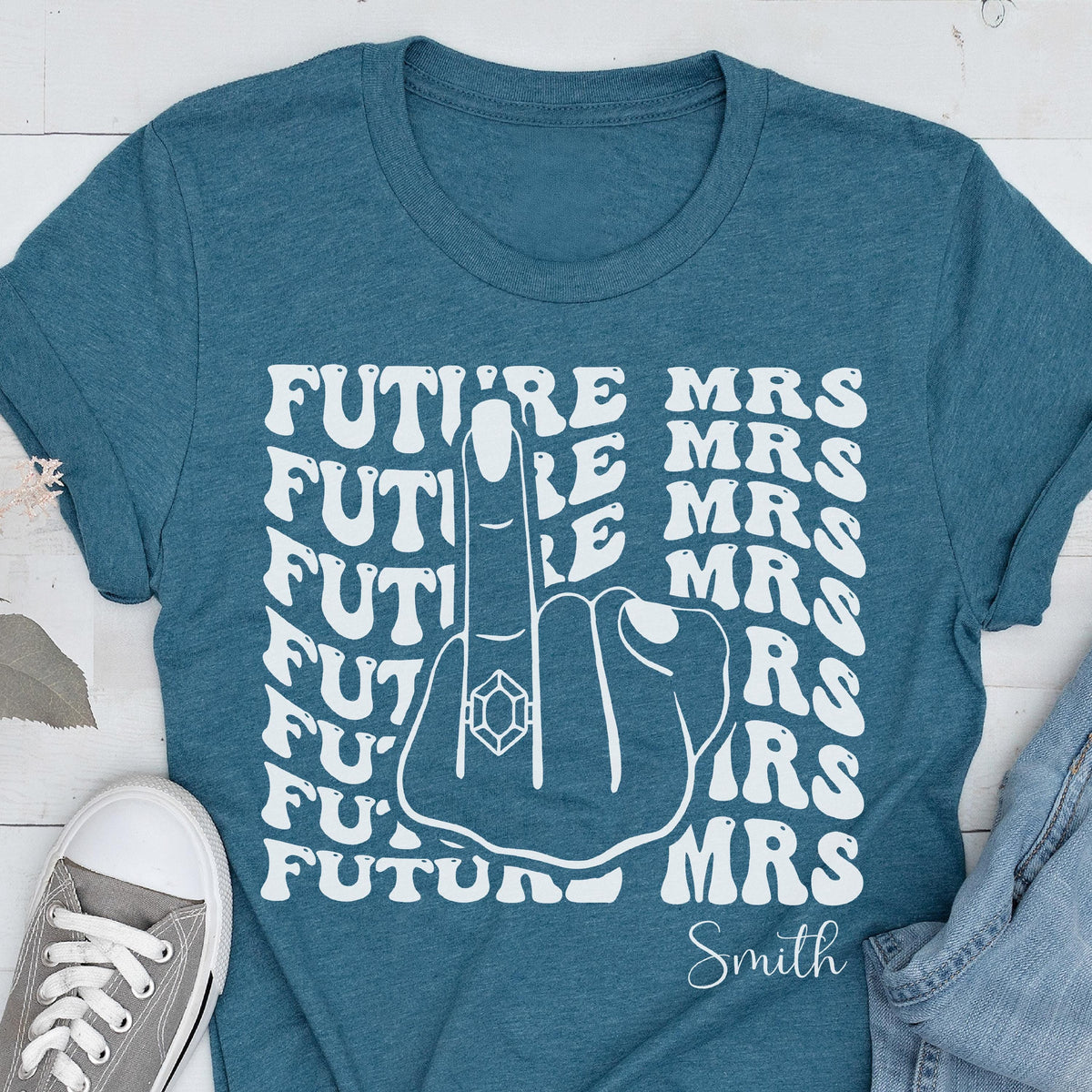 May include: A teal short-sleeved t-shirt featuring a white graphic of a hand with a ring on the middle finger. The text "FUTURE MRS" is repeated in a wavy pattern behind the hand, and the word "Smith" is printed in white script at the bottom.
