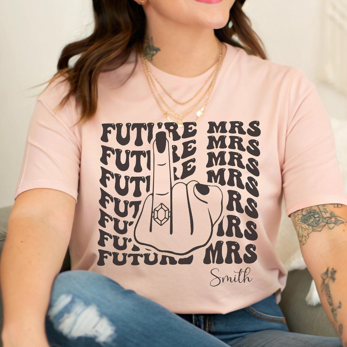 May include: A light pink t-shirt featuring a black graphic of a hand making a middle finger gesture with a ring on the finger. The text "FUTURE MRS" is repeated in a wavy pattern on both sides of the hand, with the name "Smith" printed below.