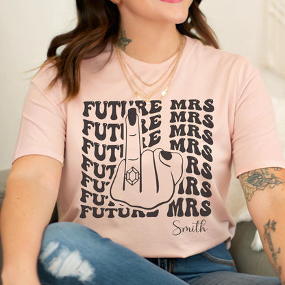 May include: A light pink t-shirt featuring a black graphic of a hand making a middle finger gesture with a ring on the finger. The text "FUTURE MRS" is repeated in a wavy pattern on both sides of the hand, with the name "Smith" printed below.