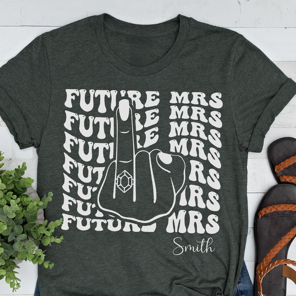 May include: A dark charcoal gray t-shirt featuring a white graphic print of a hand with a ring on the middle finger. The text "FUTURE MRS" is repeated in a wavy font pattern, and the name "Smith" is printed in a script font at the bottom.