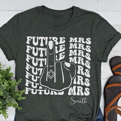May include: A dark charcoal gray t-shirt featuring a white graphic print of a hand with a ring on the middle finger. The text "FUTURE MRS" is repeated in a wavy font pattern, and the name "Smith" is printed in a script font at the bottom.