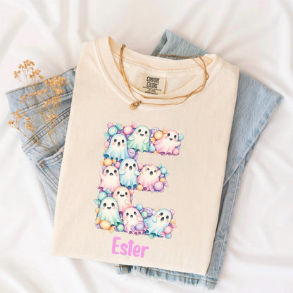 May include: A cream-colored t-shirt featuring a large letter E composed of pastel-colored ghosts and candies. Below the letter, the name Ester is printed in pink. The shirt has a Comfort Colors label at the neck and is layered over blue jeans.