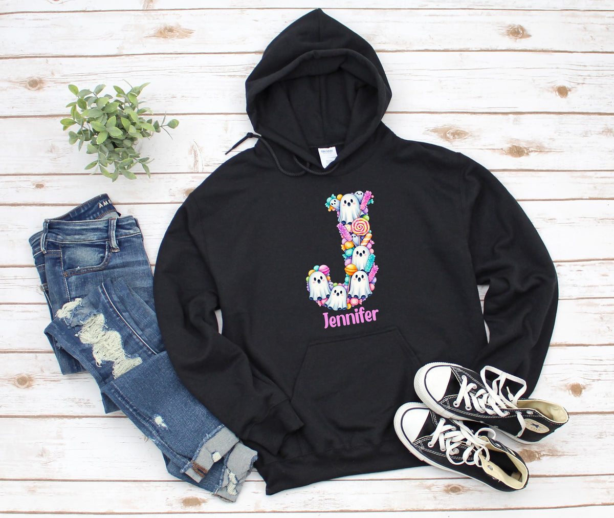May include: A black hooded sweatshirt featuring a large letter J graphic composed of white ghosts and colorful candies. The name Jennifer is printed in pink below the letter. The hoodie includes a front kangaroo pocket and matching black drawstrings.