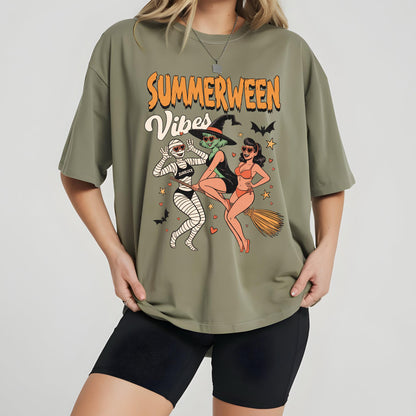 May include: Olive green t-shirt with the words "Summerween Vibes" in orange and white. The graphic features three cartoon characters: a mummy, a witch, and a woman in a swimsuit, with bats and stars.