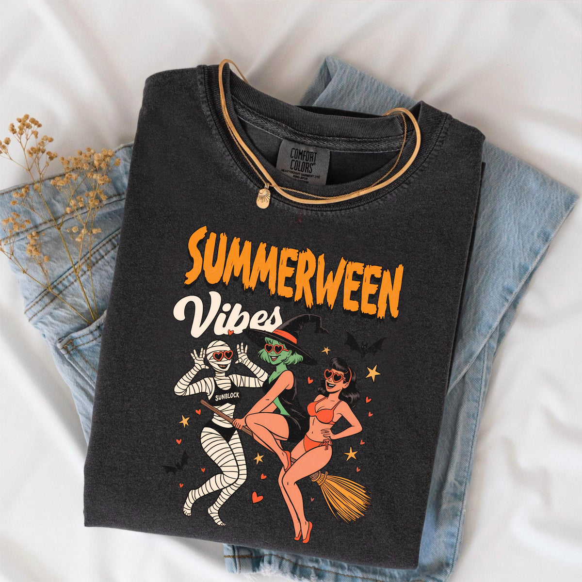May include: Dark gray t-shirt with the words "SUMMERWEEN Vibes" in orange and white. The graphic features three cartoon figures: a mummy, a witch, and a woman in a bikini. A gold necklace is draped over the shirt, which is folded on top of blue jeans.